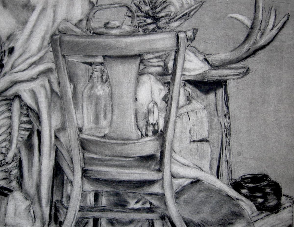 A charcoal still-life drawing featuring a wooden chair in the foreground. Behind the chair, a bottle and an animal skull are visible, creating a rustic and mysterious atmosphere. Antlers extend from the upper right side, adding to the natural and slightly eerie setting. The composition includes draped fabric and other objects, blending textures and forms in a detailed, layered arrangement.