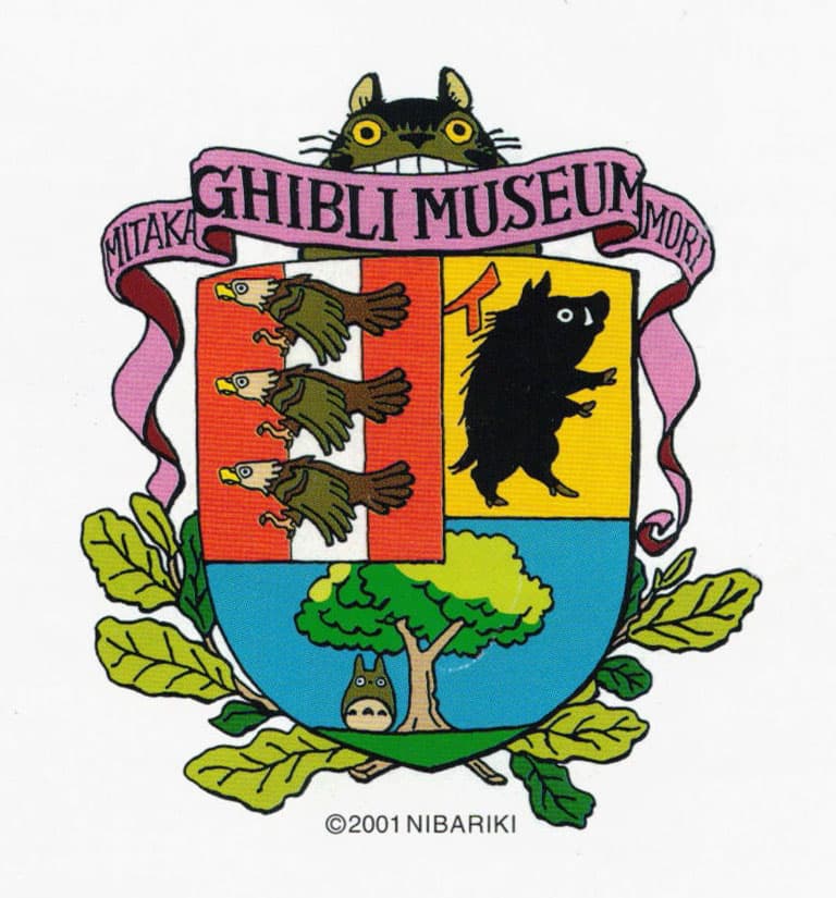 A colorful crest for the Ghibli Museum, featuring a whimsical design. The crest is divided into four quadrants: the top-left shows three flying birds, the top-right depicts a black boar, and the bottom section shows a tree with Totoro, a character from My Neighbor Totoro, peeking from below. Above the crest, a Catbus figure peers over with its wide yellow eyes. A pink banner reading "Ghibli Museum, Mitaka" wraps around the top. The crest is surrounded by green leaves, adding a natural and playful touch to the design.