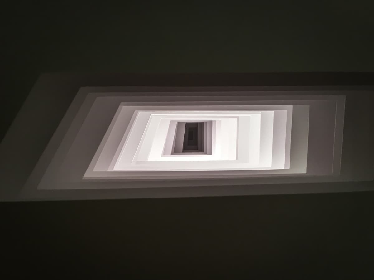View looking into a paper sculpture made of concentric square layers, creating the illusion of a spiral staircase. The soft lighting enhances the depth and geometric precision of the artwork.