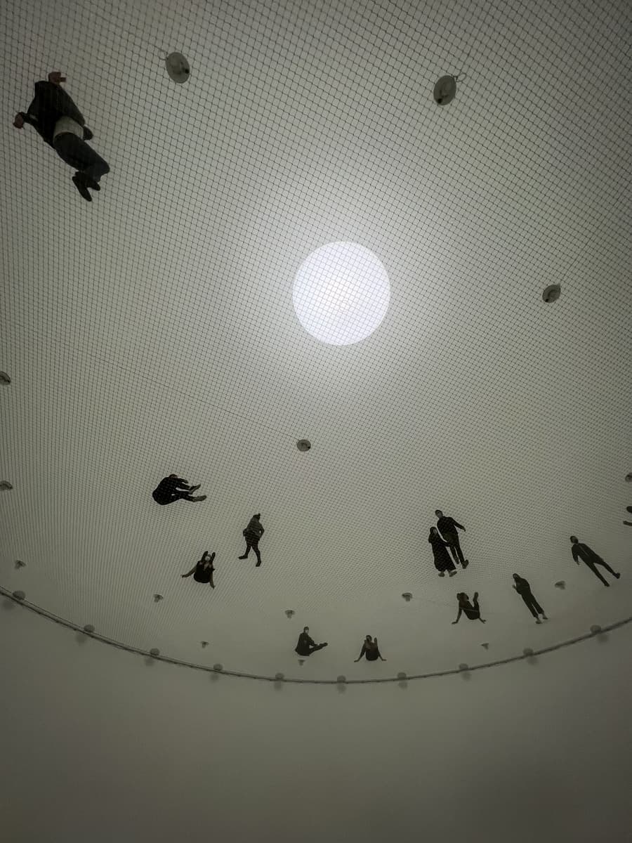 A surreal, dreamlike photograph of people suspended on a large, circular net-like structure. The figures are scattered across the surface, seen from below, as if floating or walking in midair. A bright light source in the center contrasts with the muted tones of the net and the dim space, creating an otherworldly effect.