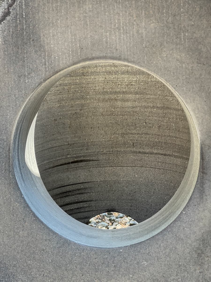 A close-up view looking down into a circular, stone opening. The interior is smooth with faint ridges, giving a sense of precision and detail in the carving. The base of the circular cavity contains small pebbles, contrasting with the smooth stone surface around it.