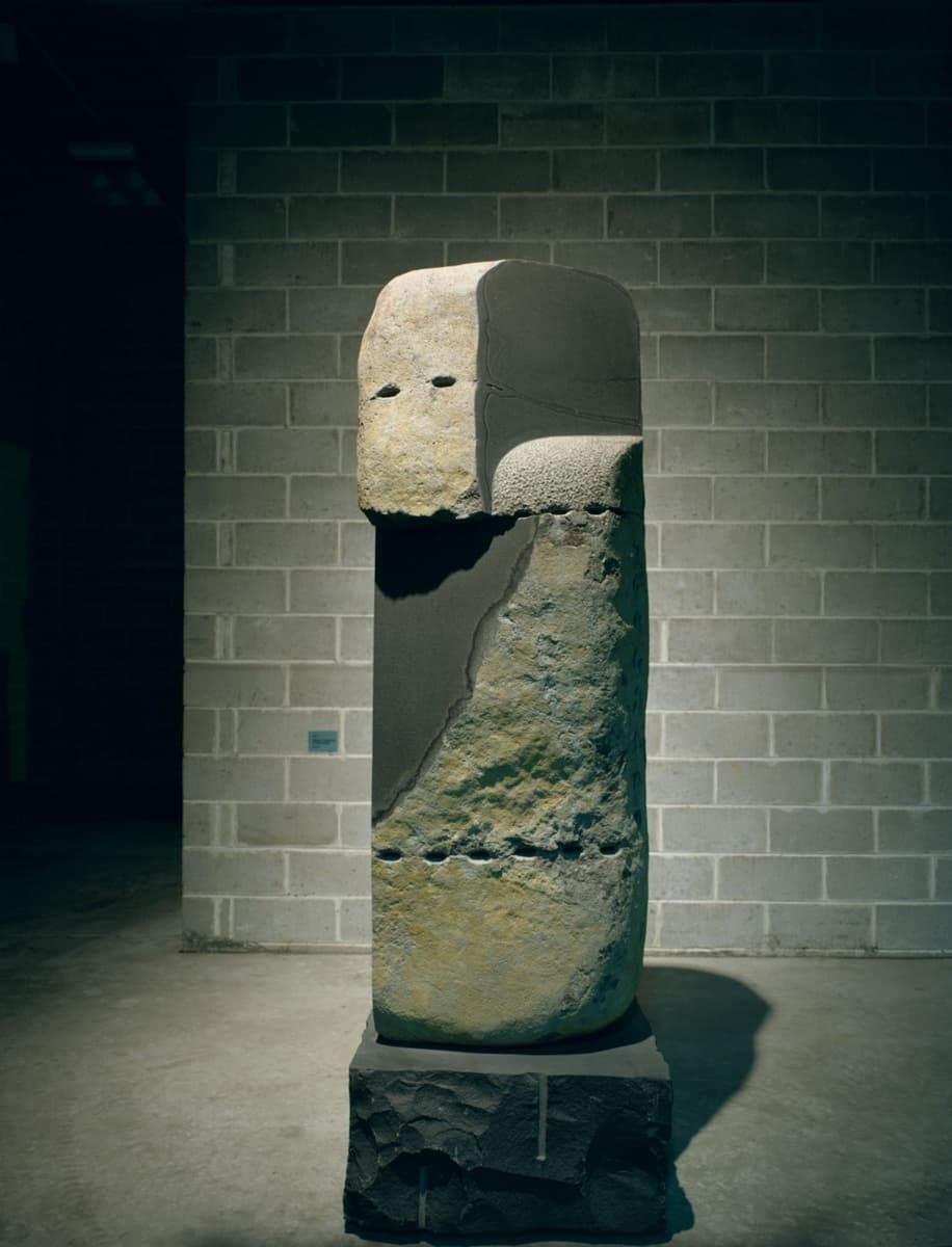 A tall, monolithic stone sculpture with a mix of rough, jagged surfaces and smoother, more polished sections. The piece appears to depict a face or abstract figure emerging from the stone, suggesting themes of transformation or release.