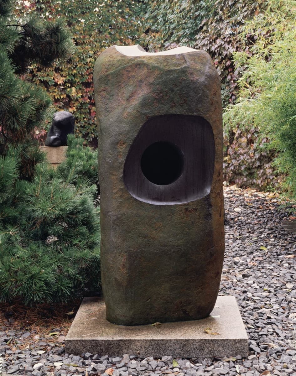 An abstract stone sculpture placed in a garden, characterized by its rough, natural texture on one side and smooth, rounded surfaces on the other. A circular void is carved into the center of the sculpture, adding a contemplative element of space within the dense material.