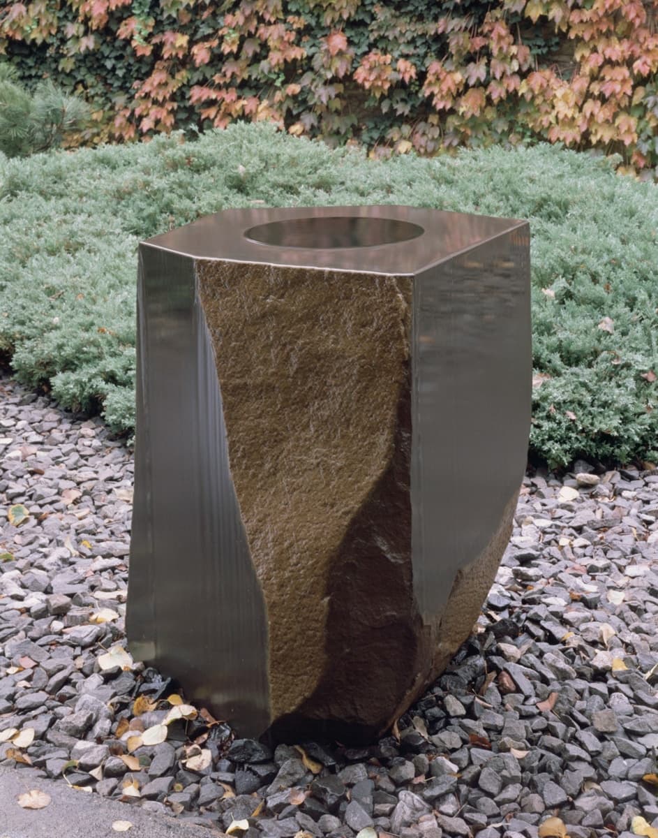 A tall, cylindrical sculpture with a rough, natural surface on the lower half and a highly polished upper section. The top has a circular opening, creating the illusion of depth. The sculpture stands outdoors on a bed of rocks, surrounded by greenery, blending the rawness of nature with refined craftsmanship.