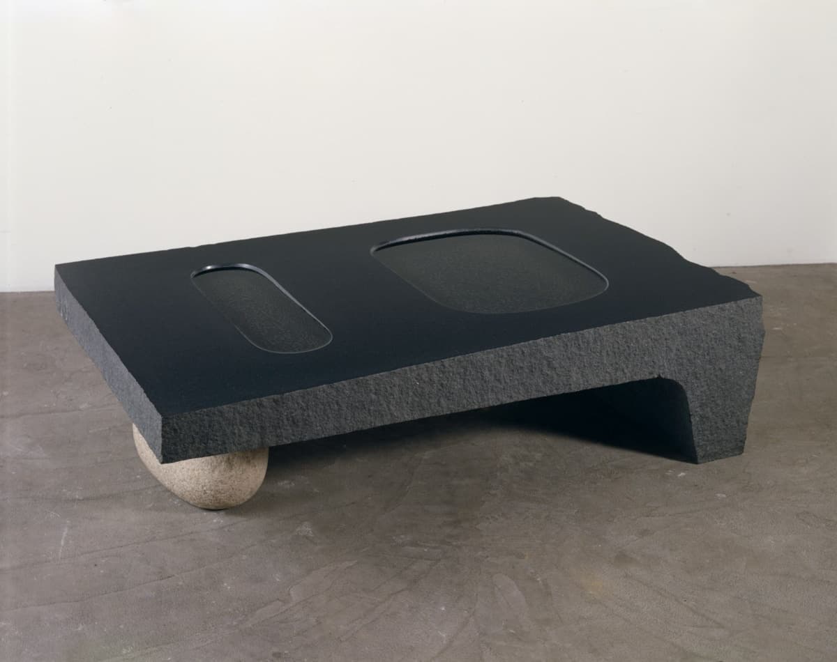 A minimalist sculpture made from a thick, flat slab of black stone, resting on a single round stone. The surface of the slab features two shallow, oval-shaped depressions. The combination of geometric precision and organic material creates a balance between nature and human intervention.