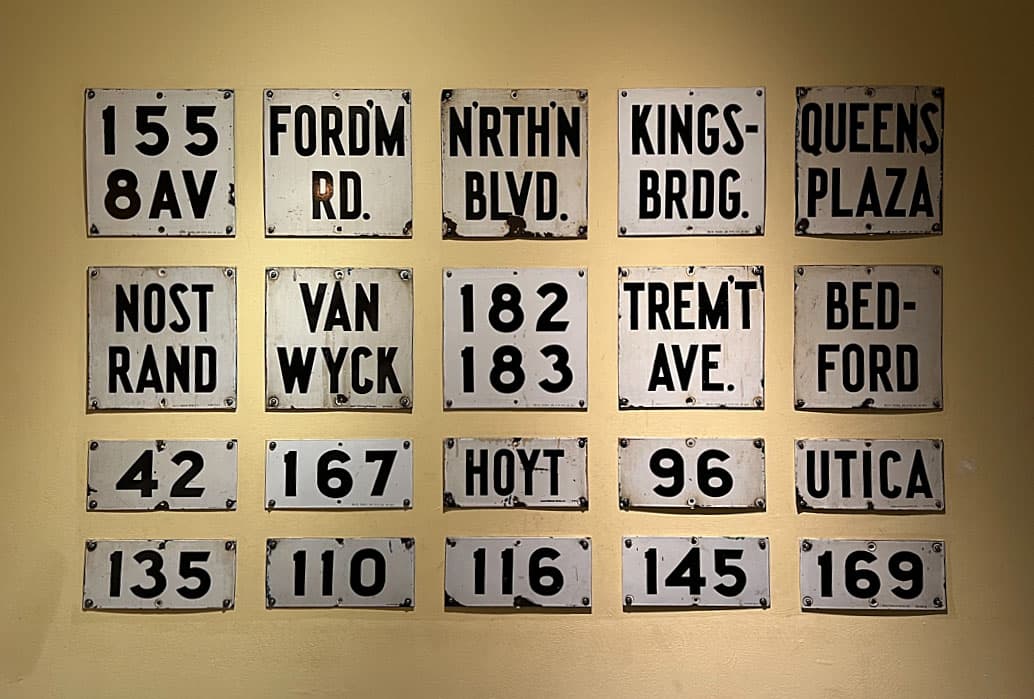 A grid of small, numbered subway signs with abbreviations for various stations and locations such as "FORDM RD" (Fordham Road), "QUEENS PLAZA," "BEDFORD," and more. The signs are weathered, emphasizing their age and historical significance.