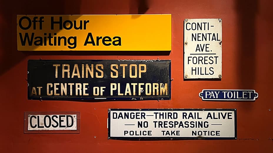 A grouping of subway-related signs, including "Off Hour Waiting Area," "Trains Stop at Centre of Platform," and a safety warning about the third rail. The signs reflect different periods and design styles, adding to the vintage atmosphere.