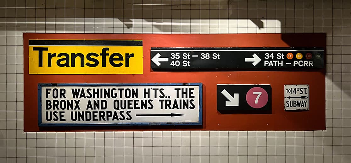 A set of subway transfer signs, with a prominent yellow "Transfer" sign alongside directional arrows for 34th Street and other destinations. The signs guide passengers for connections to The Bronx, Queens, and Washington Heights.