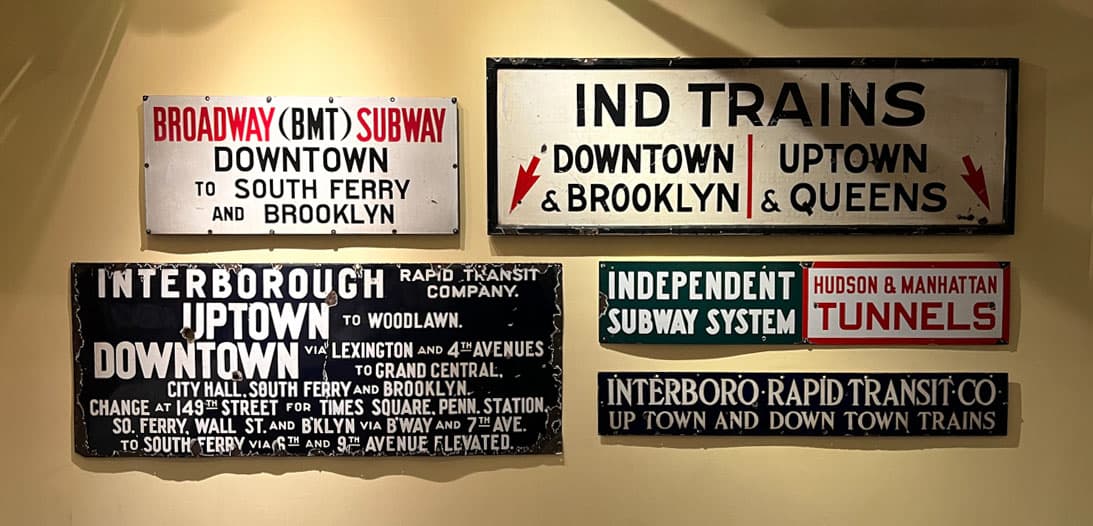 A display of various old subway route signs, including one for "Broadway (BMT) Subway" and directions to South Ferry and Brooklyn. Additional signs include "IND Trains Downtown & Brooklyn" and "Independent Subway System," all using distinctive fonts and arrow graphics.