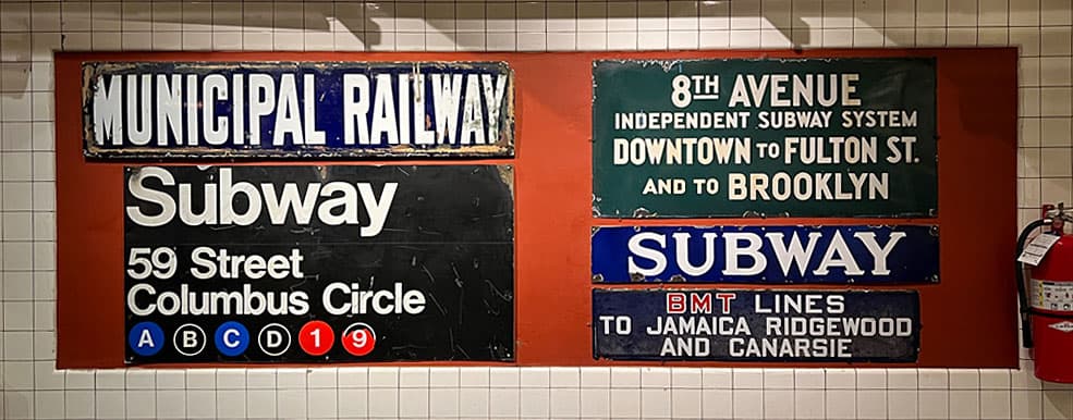 A collection of vintage subway signs, including "Municipal Railway," "Subway" for the 59th Street Columbus Circle station, and signs for the 8th Avenue Independent Subway System. The signs feature bold fonts and iconic colors, such as black, white, and green.
