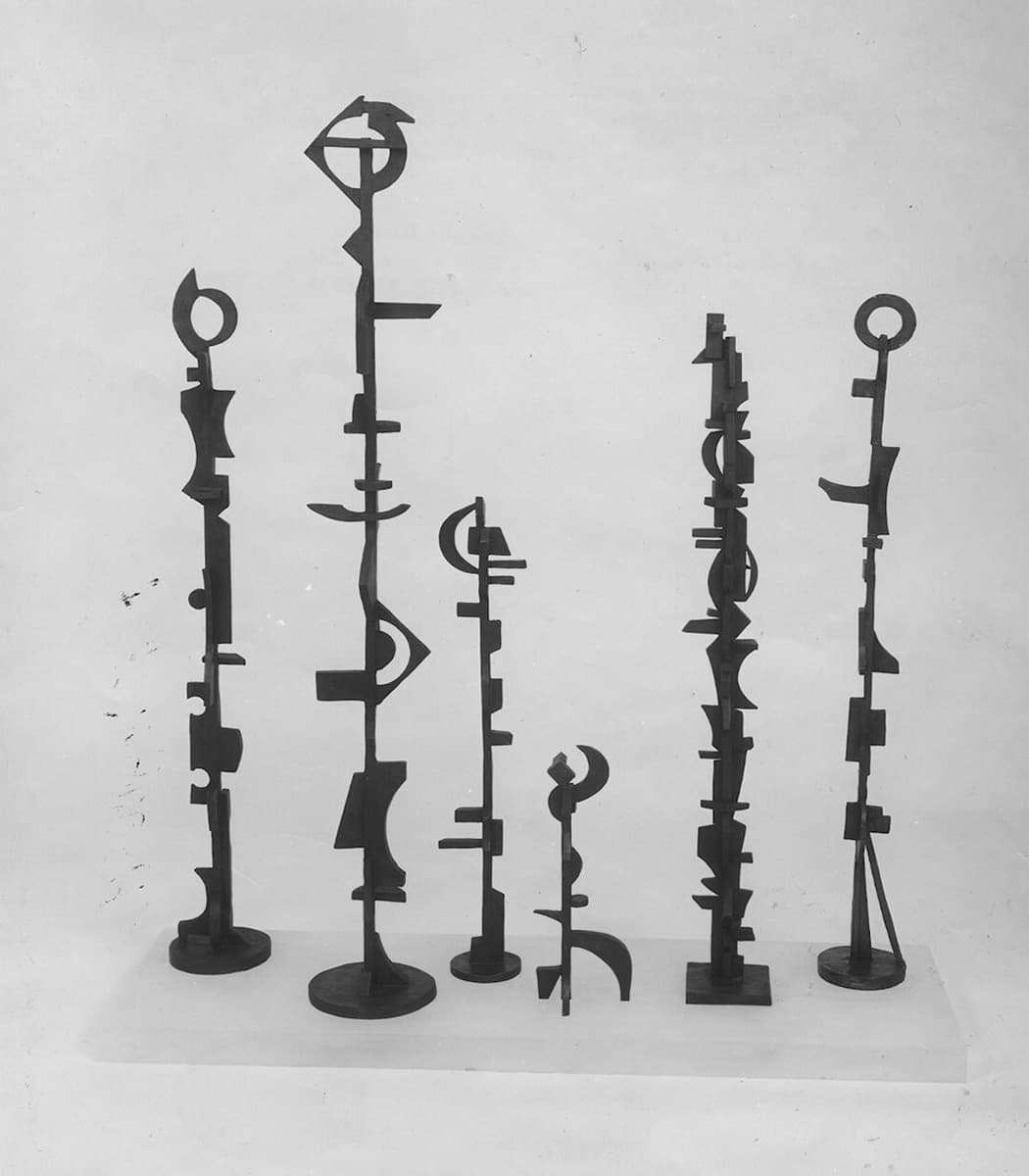 A group of six slender, abstract metal sculptures of varying heights, with geometric and organic shapes, displayed against a plain background, resembling totems or ancient symbols.