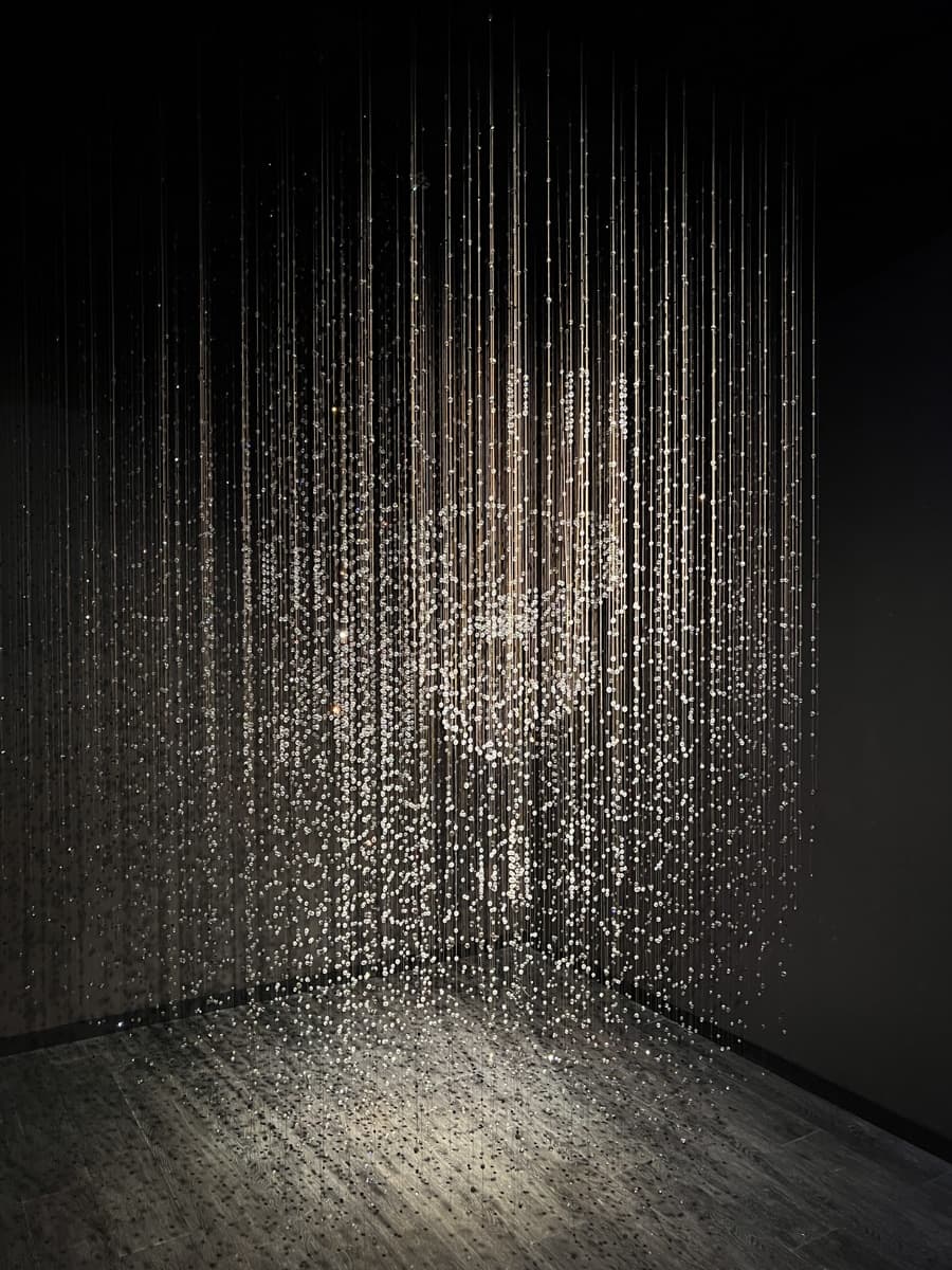 An image of an art installation made of hanging strings with small reflective or illuminated beads. The beads catch the light, creating a shimmering effect in a dark room, almost like suspended droplets of water or a cascading wave of light. The installation gives a sense of movement and flow, creating a magical, ethereal ambiance.