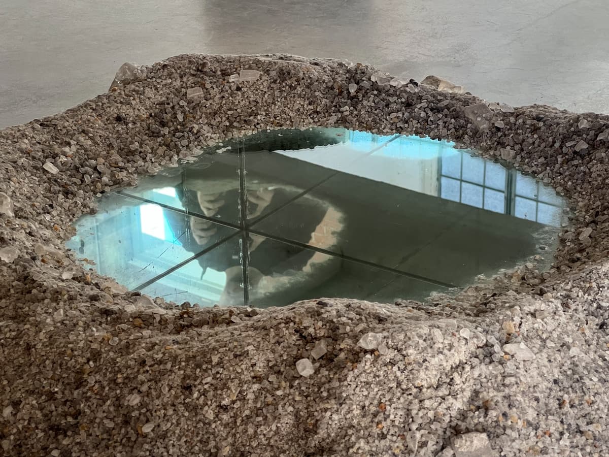 This image showcases what looks like a pool of water within a rough stone or gravel formation. The pool's surface reflects the surroundings, including a figure and a bright window, creating a captivating interplay between reality and reflection. The stillness of the water contrasts with the rugged edges of the stone, suggesting a meeting between natural and constructed environments.