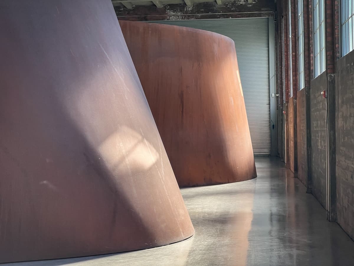 This image features massive, rust-colored curved structures in a spacious industrial setting. The light casts soft shadows on the surfaces, emphasizing their scale and solidity. These abstract forms give off a monumental, almost architectural presence, inviting viewers to consider the relationship between space, form, and light.
