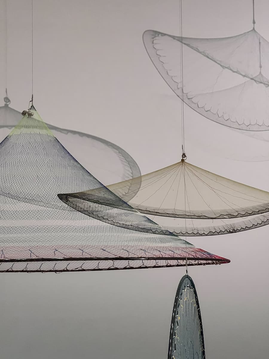 This image features a delicate art installation of net-like structures or canopies, each suspended from the ceiling. They seem to float in the air, with translucent layers and soft, subtle colors. The overlapping shadows on the wall create a feeling of depth, adding to the sense of weightlessness and gentle movement in the space.