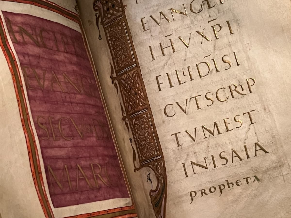 A close-up photograph of an illuminated manuscript, featuring Latin text in gold leaf. On the left page, the text is faded and written on a purple background with decorative red borders. The right page has large, clearly visible gold letters with ornate, intricate decoration along the vertical margin. The craftsmanship emphasizes the luxurious and historical nature of the manuscript, with the text likely from a religious or classical source.