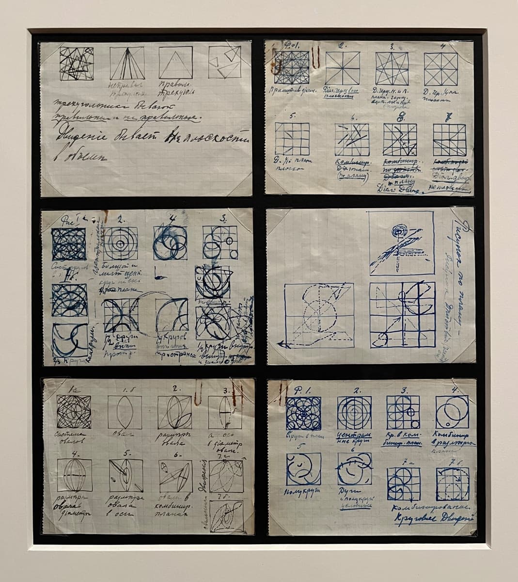 A collection of graph paper sheets displayed in a frame, featuring intricate hand-drawn geometrical sketches in blue ink. The drawings include various circles, squares, and intersecting lines, accompanied by handwritten notes and labels in a mix of cursive and printed text. The sketches appear to study geometric patterns and mathematical principles, meticulously drawn and annotated.