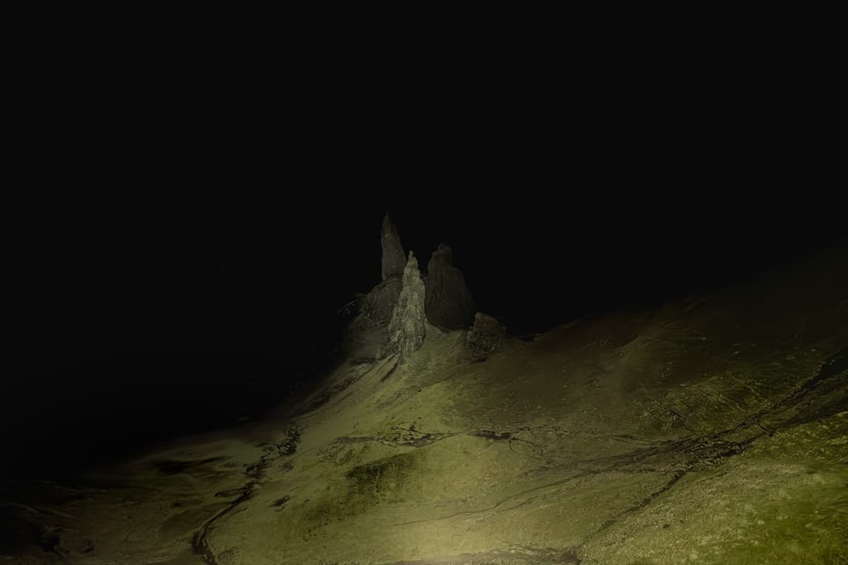 A dramatic nighttime landscape of jagged rock formations emerging from a gently sloping hill. The dim light creates a mysterious mood, casting shadows over the rocky peaks and the barren ground beneath, all set against a backdrop of an almost completely black sky.