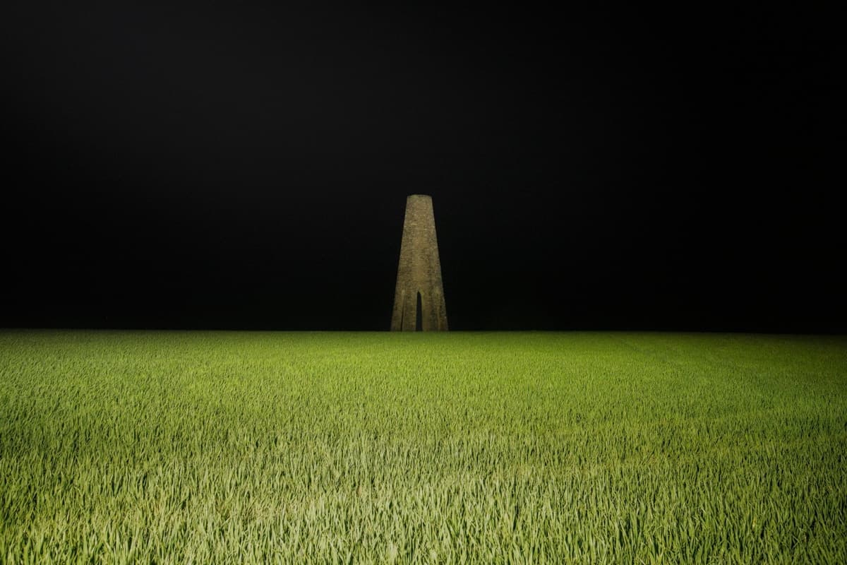A surreal night photograph showing a tall, narrow stone structure rising from a vast green field, illuminated by soft lighting. The stark contrast between the brightly lit grass and the pitch-black sky creates an eerie, otherworldly atmosphere, with the solitary tower-like structure as the central focus.