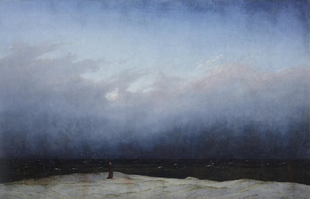 A lone monk stands at the edge of a bleak, wind-swept shore. Above him looms an enormous sky, heavy with mist and sea spray. The figure is dwarfed by the elemental vastness around him.