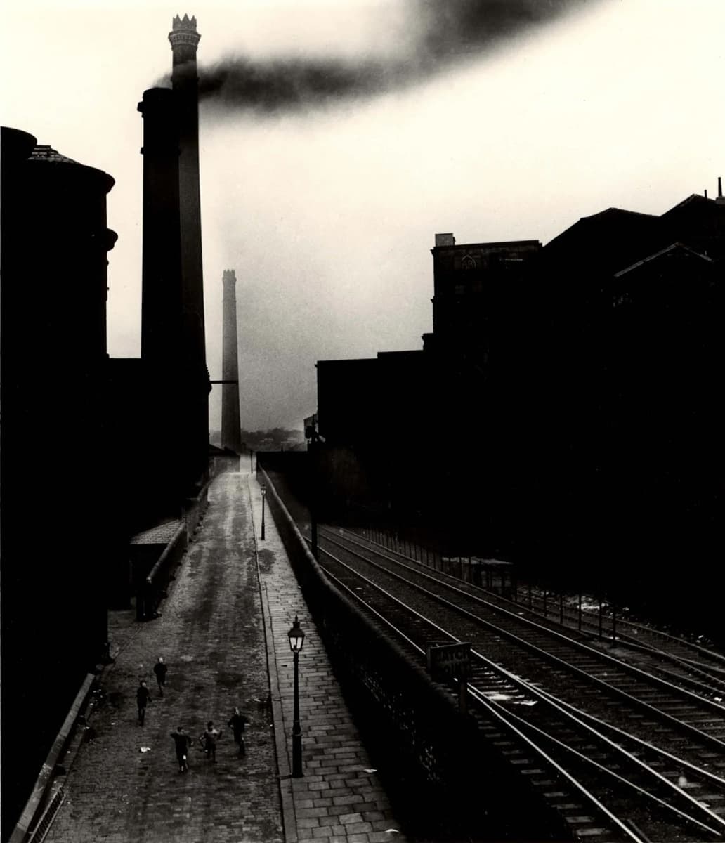 A black-and-white photograph of an industrial street scene, likely from mid-20th century Britain. A group of children walk along a cobbled road flanked by looming factory buildings. Two tall smokestacks dominate the skyline, with one releasing a thick plume of black smoke. Railway tracks run parallel to the road on the right side, bordered by a stone wall. The image is atmospheric, with heavy contrast between the smoke-darkened sky and the silhouettes of industrial structures.