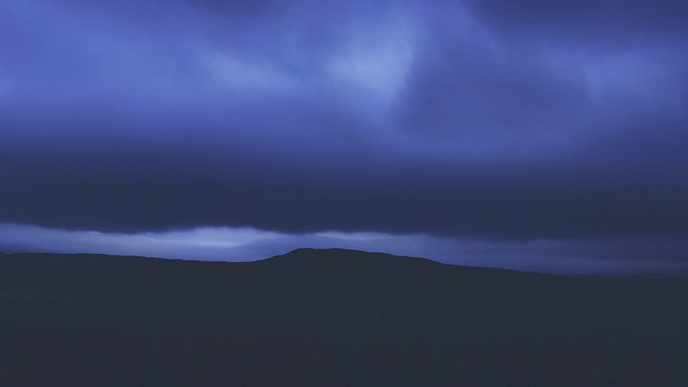Clouds cascade over a dark mountain ridge in a dramatic display, creating the appearance of misty waterfalls beneath a pale blue sky.