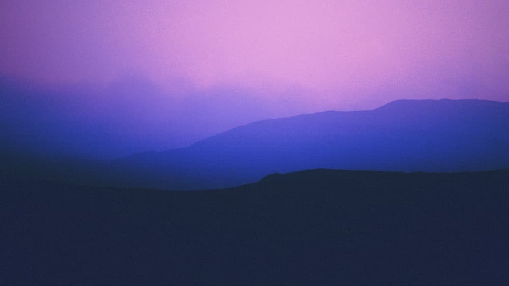 Layered mountain ridges fade into a misty horizon, bathed in ethereal shades of purple and blue beneath a glowing pink sky.