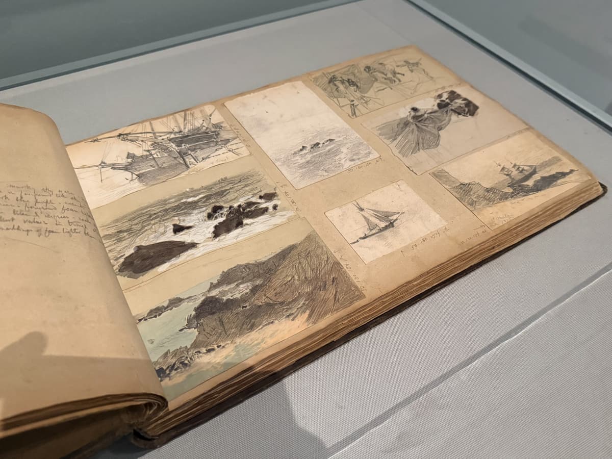 A worn sketchbook lies open in a display case, revealing multiple detailed pencil and ink studies. The pages contain seascapes, sailing ships, rocky coastlines, and figures in motion, showing the artist’s observational process and travel documentation.