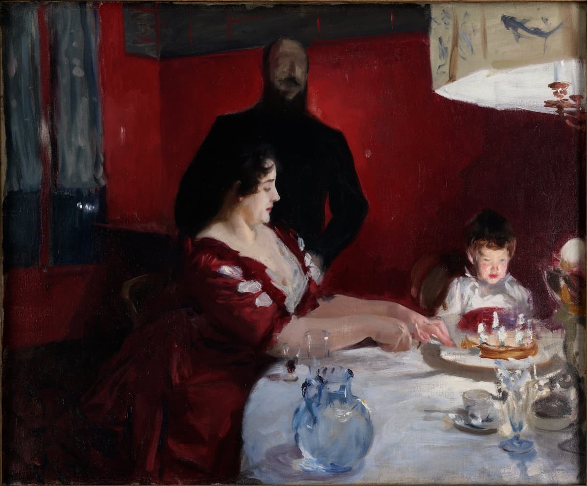 A richly lit painting of a family gathered around a white-clothed dining table. A woman in a deep red dress cuts a cake for a child sitting beside her, while a shadowy male figure stands behind them. The red walls and soft lamplight create a dramatic contrast of light and shadow, emphasizing intimacy and warmth.