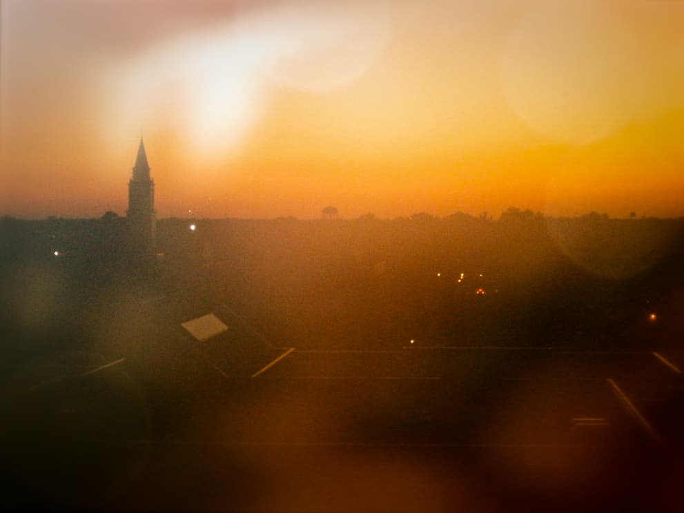 This photograph captures a hazy, golden sunset over a town or campus. The sun is low on the horizon, casting a warm orange and yellow glow across the sky. The silhouette of a tall spire is visible against the fading light, and faint lights from buildings or streets can be seen in the distance. The image has a soft, blurred quality, with lens flare effects adding a dreamy, ethereal atmosphere. The combination of the warm sunset and the misty, out-of-focus foreground creates a tranquil, almost surreal mood.