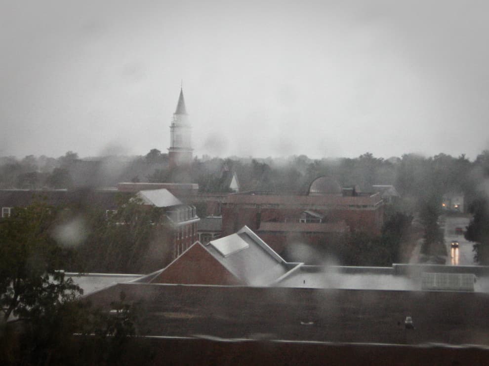 This photograph shows a rainy, overcast day with a view of a town or campus. The scene is blurred by raindrops on the window, adding to the dreary and misty atmosphere. In the background, a tall spire is visible, partially obscured by the rain and fog, with rooftops and trees in the foreground. The wet streets reflect the dim light of the day, and the overall mood feels calm yet somber, capturing the essence of a stormy or rainy afternoon. The muted colors and softened outlines enhance the feeling of rain-soaked tranquility.