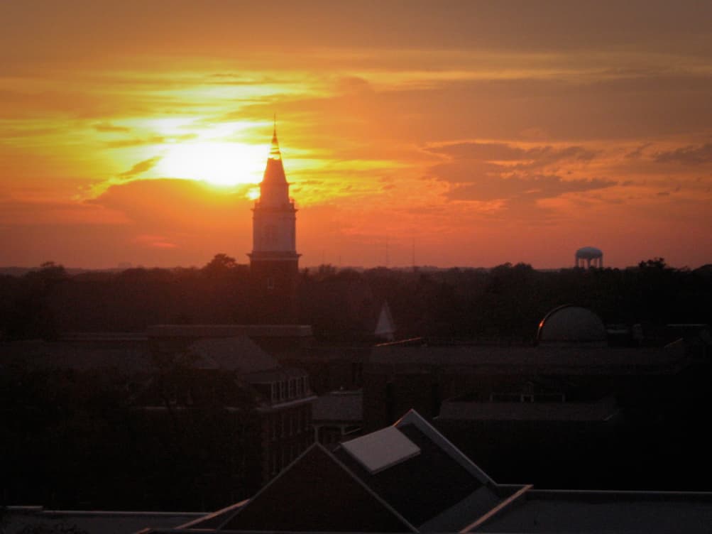 This photograph captures a stunning sunset over a town or cityscape. The sun, glowing in deep orange and yellow hues, is partially hidden behind clouds, casting a warm light over the scene. Silhouetted against the sunset is a tall, prominent steeple, likely belonging to a church or historic building, which stands out as the focal point of the image. In the distance, a water tower can also be seen, along with trees and rooftops, all in shadow due to the fading light. The sky is filled with soft, glowing clouds, adding to the peaceful and dramatic atmosphere.