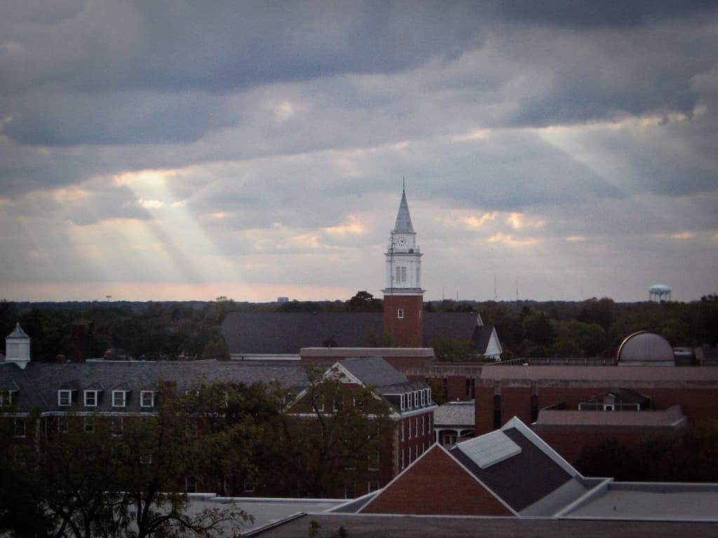 This photograph shows a cloudy sky over a town or campus with dramatic beams of sunlight breaking through the clouds, creating a serene and almost ethereal atmosphere. In the center of the image, a tall brick tower with a white steeple and clock stands prominently. The surrounding buildings, trees, and rooftops are bathed in soft light from the breaks in the clouds. In the distance, a water tower and other structures are visible. The combination of the overcast sky with rays of sunlight adds a peaceful and contemplative mood to the scene.