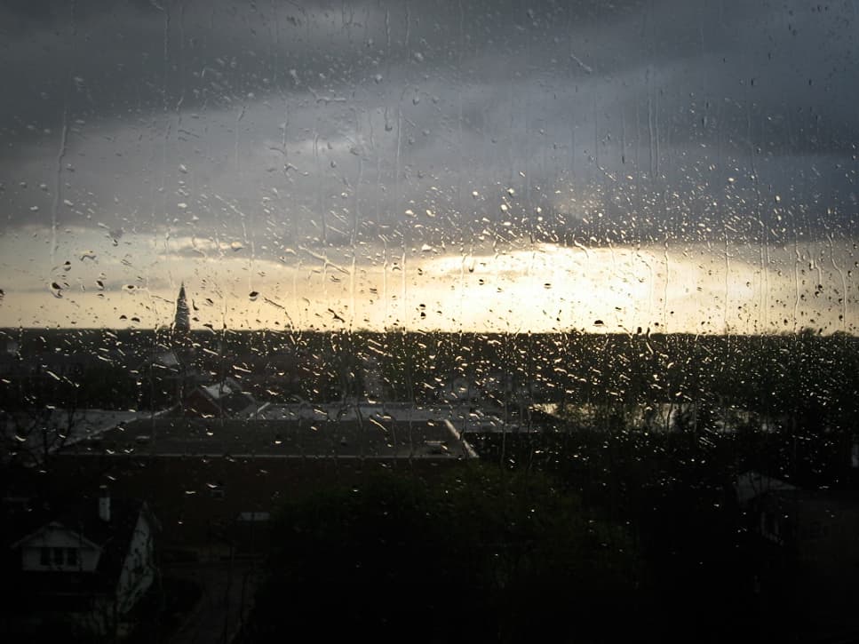 This photograph captures a rainy day, with raindrops covering a window, through which the distant landscape is visible. The sky is overcast, and the sun is low on the horizon, casting a faint glow and illuminating the rain-splattered window. A silhouette of a tall spire can be seen in the background, surrounded by rooftops and trees. The contrast between the gloomy weather and the soft light breaking through the clouds adds a melancholic yet peaceful feeling to the scene. The focus on the raindrops creates a textured and immersive effect.