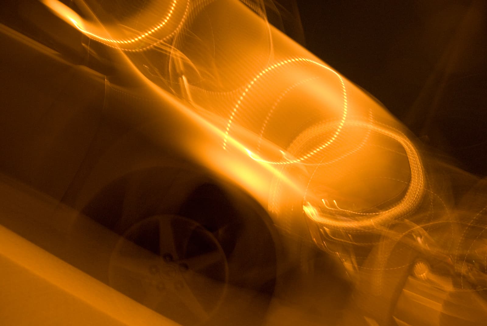 This abstract photograph features swirling golden light trails that twist and curve across the image, creating a sense of dynamic movement. In the background, the blurred outline of a car's wheel and body can be faintly seen, though the primary focus remains on the light. The circular patterns of the light create a mesmerizing effect, blending with the warm glow of the background. The motion blur gives the image an ethereal and dreamlike quality, evoking a sense of speed and fluidity, as if capturing the energy of a fleeting moment.
