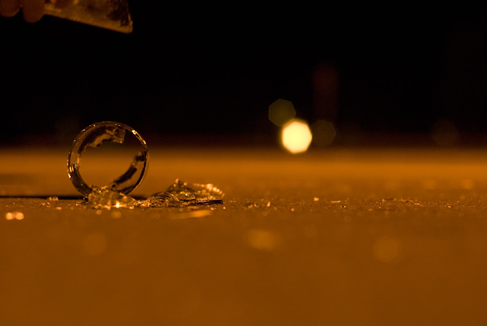 This close-up photograph shows broken glass scattered on the ground, with a circular fragment standing upright among the shattered pieces. The scene is bathed in warm, golden lighting, with soft bokeh lights in the background, adding a sense of depth and focus to the image. The contrast between the sharp edges of the broken glass and the soft, blurred background creates a dramatic and contemplative mood, evoking feelings of fragility and tension. The low angle and focus on the glass emphasize the moment of stillness after something has been broken.