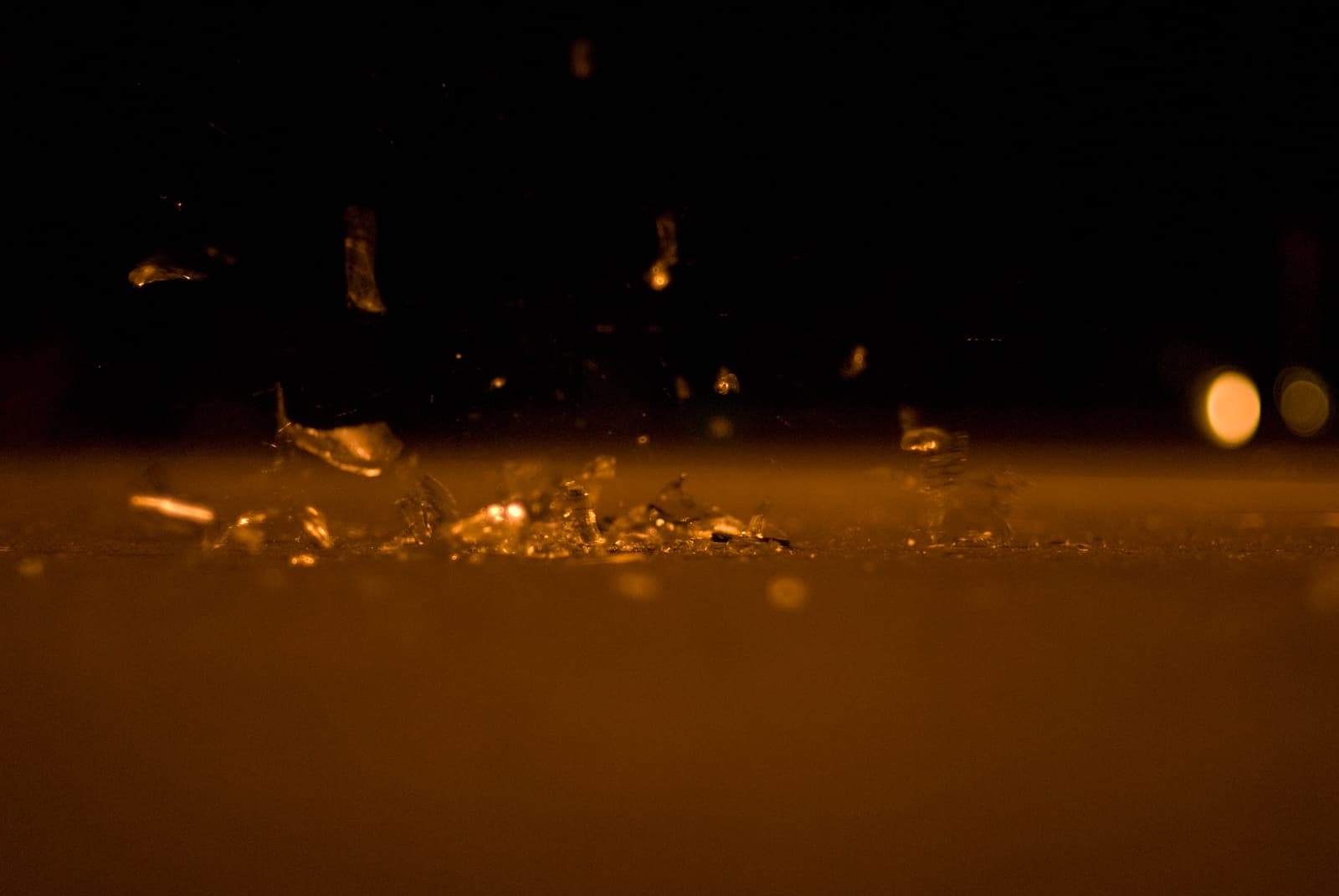 This close-up photograph captures shattered glass pieces scattered across the ground, with a few fragments suspended in mid-air, as if just recently broken. The scene is bathed in warm, golden lighting, creating a striking contrast between the sharp glass shards and the smooth, blurred background. The faint bokeh lights in the distance add a sense of depth and atmosphere. The image conveys a moment of action and fragility, emphasizing the delicate nature of the glass and the quiet stillness after a sudden break.