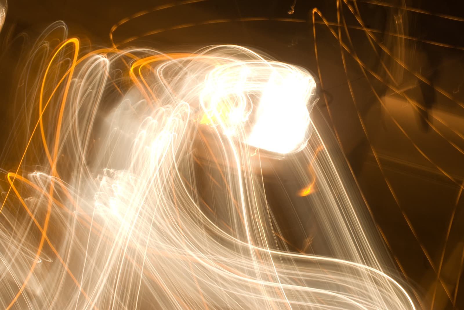 This abstract photograph features bright white and orange light trails, creating fluid and energetic patterns across the image. The lights twist and curve in sweeping motions, forming looping and intersecting lines. The vivid white light at the center radiates outward, while the surrounding orange lines add depth and contrast. The overall effect is one of intense movement and dynamic energy, as though capturing the essence of motion in a single frame. The image's long-exposure style gives it a sense of flow and fluidity, reminiscent of illuminated pathways or light waves in a dreamlike setting.