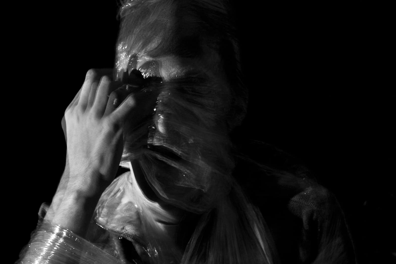 This black-and-white photograph features a person wrapped in a transparent plastic film, obscuring their face and upper body. The light reflects off the plastic, creating a shiny, distorted texture. The figure's hand is pulling at the film near their face, as if attempting to free themselves or make a space to breathe. The combination of the plastic and the shadowy lighting adds a sense of tension and confinement to the image, evoking feelings of suffocation or entrapment. The dark background enhances the isolation of the figure, giving the photograph an eerie, unsettling mood.