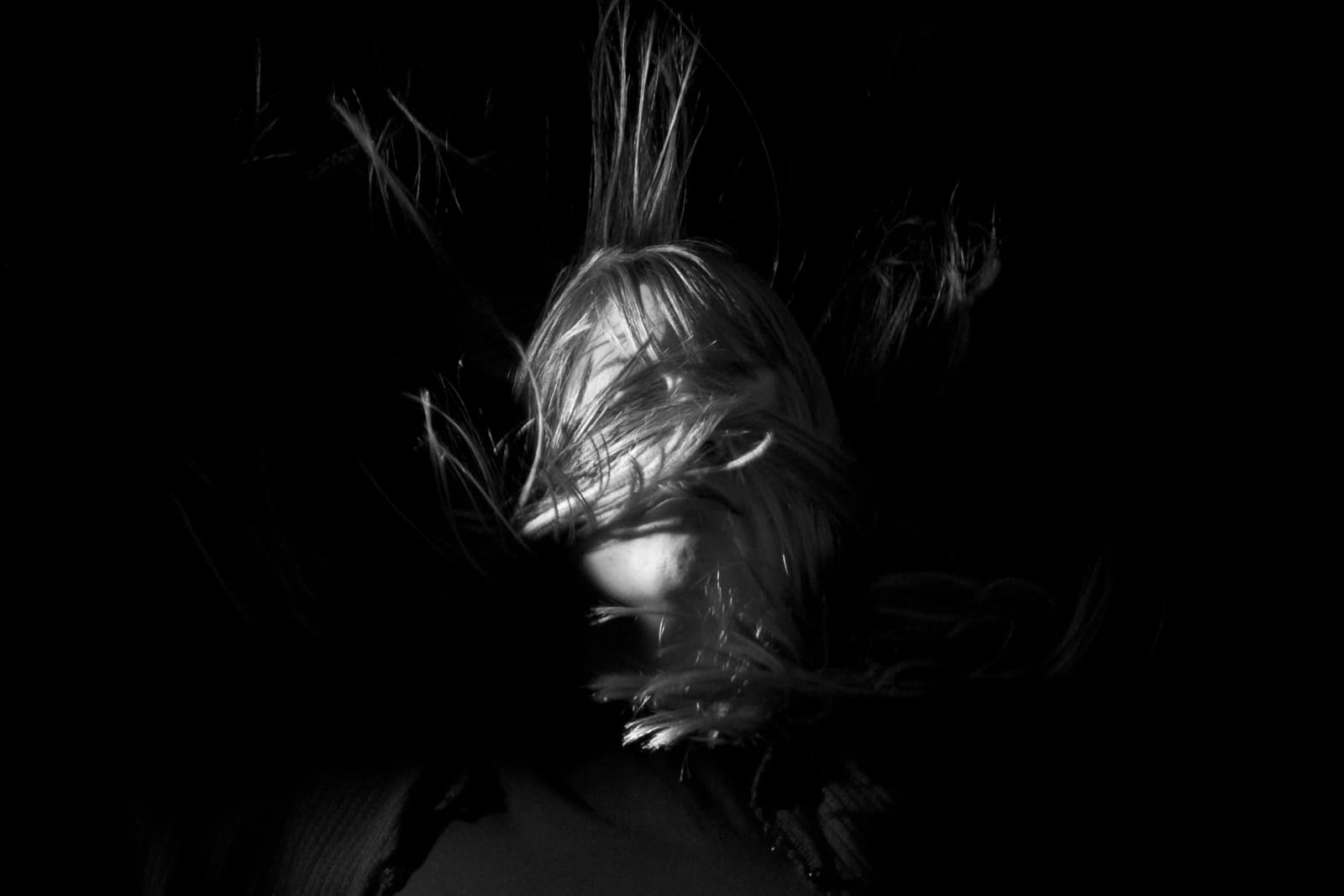 This black-and-white photograph shows a person with their face obscured by strands of hair, which are illuminated in sharp contrast against a dark background. The hair appears to be floating or blown by wind, adding a sense of motion and mystery to the image. Only part of the face is visible, emphasizing the eyes and nose, while the rest is hidden beneath the tangled hair. The play of light and shadow creates a dramatic, almost haunting atmosphere, as the figure seems to emerge from or be consumed by the darkness. The image evokes feelings of vulnerability, disorientation, or isolation.