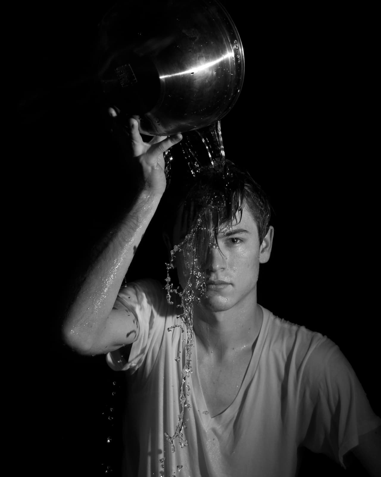 This black-and-white photograph shows a young man pouring water over his head from a large metal bowl. The water cascades down his face and body in sharp detail, with droplets and streams frozen mid-motion. His expression is calm and serious, and his wet hair hangs over part of his face. He wears a simple, wet t-shirt, which clings to his body as the water continues to flow. The dark background isolates the subject, emphasizing the contrast between the water, his skin, and the reflective bowl. The image captures a sense of stillness and focus, despite the movement of the water.