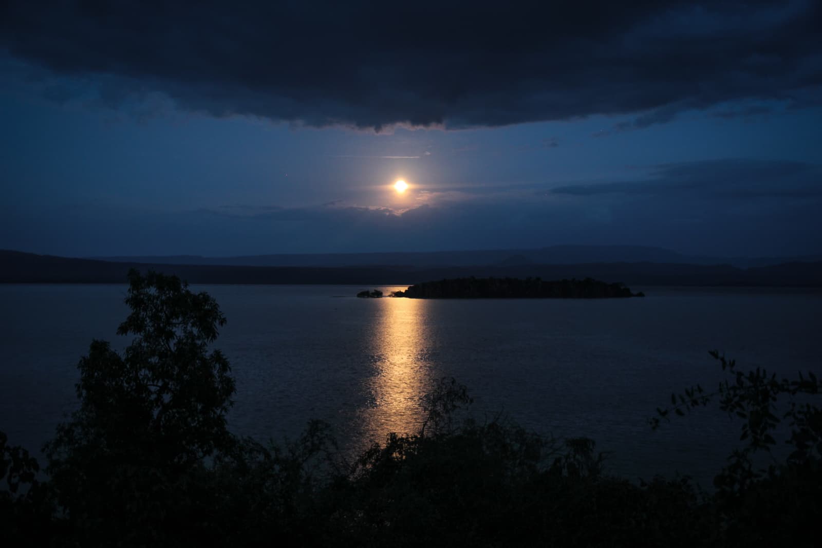 A serene nighttime scene over a body of water, with the full moon rising above the horizon. The moon casts a reflective light on the water, creating a glowing path that stretches toward the viewer. Silhouettes of trees are visible in the foreground, and a small island can be seen in the distance. The sky is mostly clear with some clouds, adding to the tranquil, almost ethereal atmosphere of the image.
