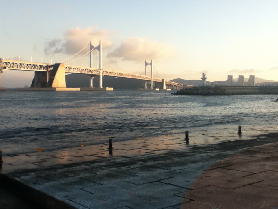 A scenic view of a large suspension bridge extending across a body of water during what appears to be either sunrise or sunset. The sky is mostly clear, with a few clouds illuminated by soft, golden light. A lighthouse stands on a small rocky peninsula near the shore, and in the distance, modern high-rise buildings are visible against a backdrop of low mountains. The foreground features a paved walkway along the water's edge, adding a serene and reflective quality to the image.