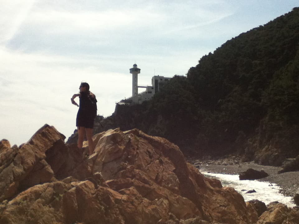 A person stands confidently on a large, jagged rock formation overlooking the sea. They are dressed in dark clothing, holding a backpack, and appear to be looking off into the distance. Behind them, on a cliffside covered in dense greenery, a tall lighthouse and a few nearby buildings are visible, perched above a rocky shoreline. The sky above is mostly clear with soft clouds, adding to the tranquil, yet adventurous atmosphere of the scene.