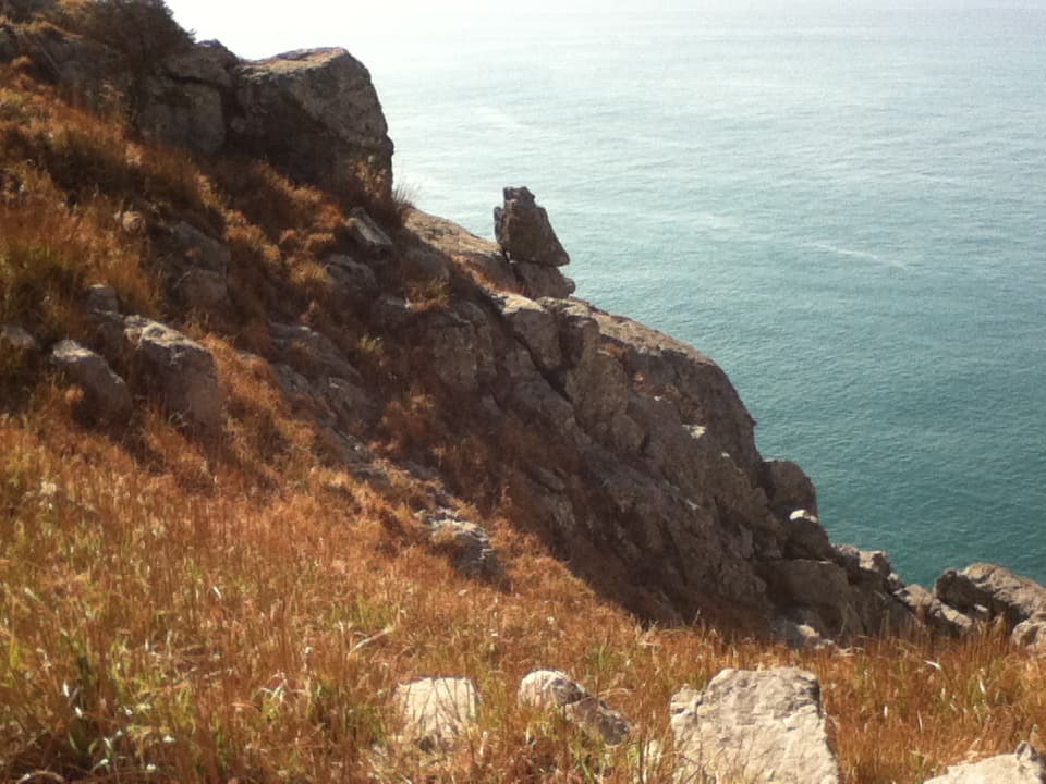 A rugged cliffside overlooking a calm ocean, with large rocks and dry, golden grass covering the slope. A solitary, balanced rock formation stands prominently on the edge of the cliff, creating an intriguing natural feature. The ocean below is a tranquil blue, extending into the horizon, offering a sense of isolation and peacefulness. The combination of the earthy tones of the rocky landscape and the serene ocean creates a striking contrast.