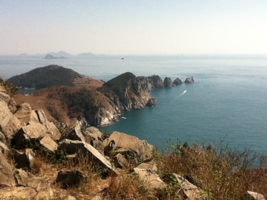 A breathtaking view from a rocky cliff overlooking a calm blue sea. The coastline below features rugged, jagged rock formations jutting out into the water, with a small speedboat leaving a trail as it moves through the ocean. In the distance, several smaller, tree-covered islands are visible, fading into the hazy horizon. The rocky terrain in the foreground is dotted with patches of dry grass, adding a sense of height and natural beauty to the scene. The sky is clear and expansive, creating a peaceful and serene atmosphere.