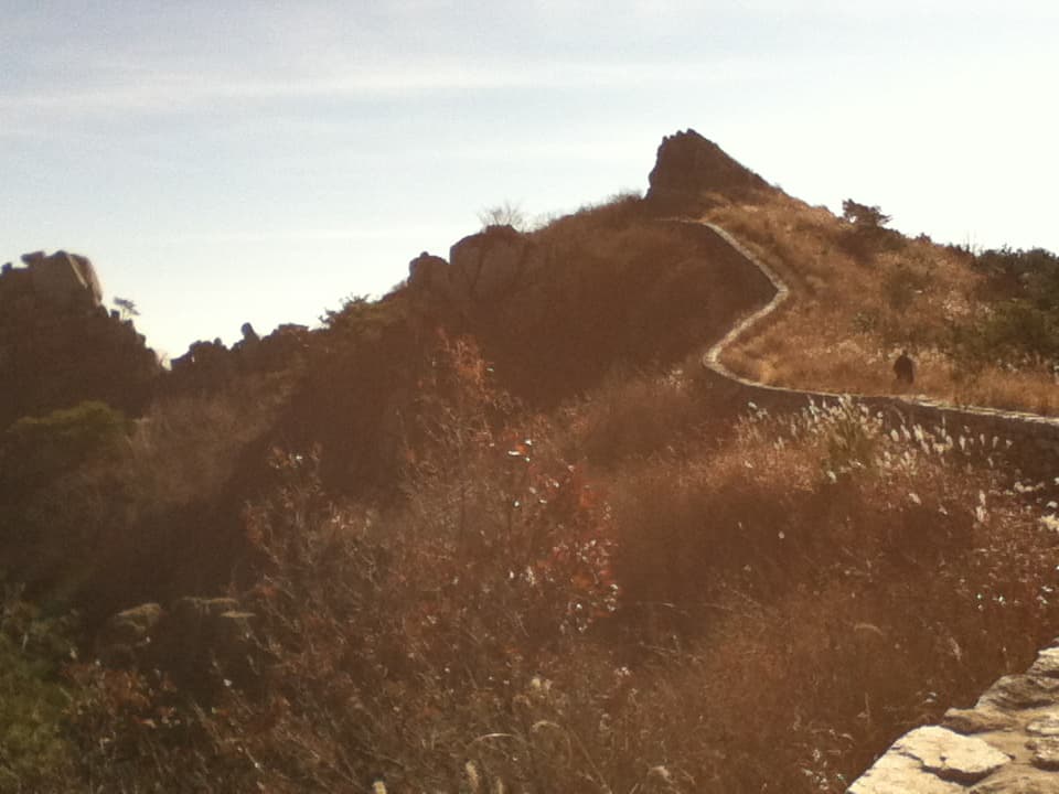 A narrow, winding stone path curves along a grassy ridge toward a rocky hilltop in the distance. The path is bordered by tall, dry grasses and low shrubs, with sunlight creating a soft, warm glow over the landscape. A lone figure walks along the path, adding a sense of solitude and peaceful exploration to the scene. The rugged terrain and jagged rocks in the background emphasize the natural beauty and tranquility of the area.