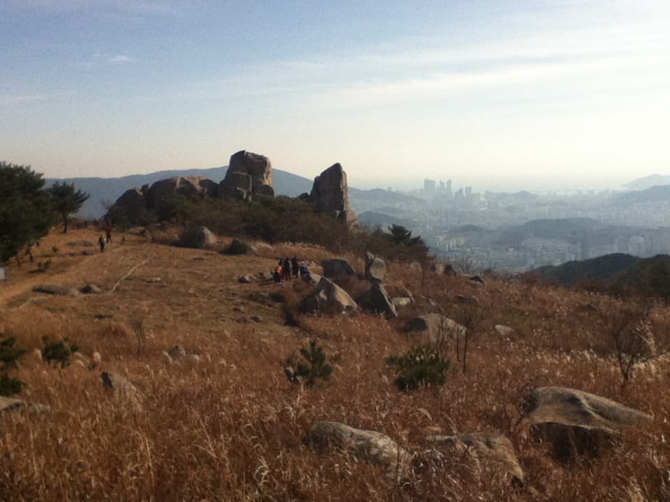 A scenic hilltop with large rock formations in the middle distance, standing prominently against the sky. The grassy terrain is dotted with rocks and small trees, with people walking along a path that leads toward the rocks. In the background, a cityscape fades into the horizon, partially obscured by mist, giving a sense of both natural beauty and urban proximity. The sunlight casts a warm, golden hue over the landscape, creating a peaceful and inviting atmosphere.