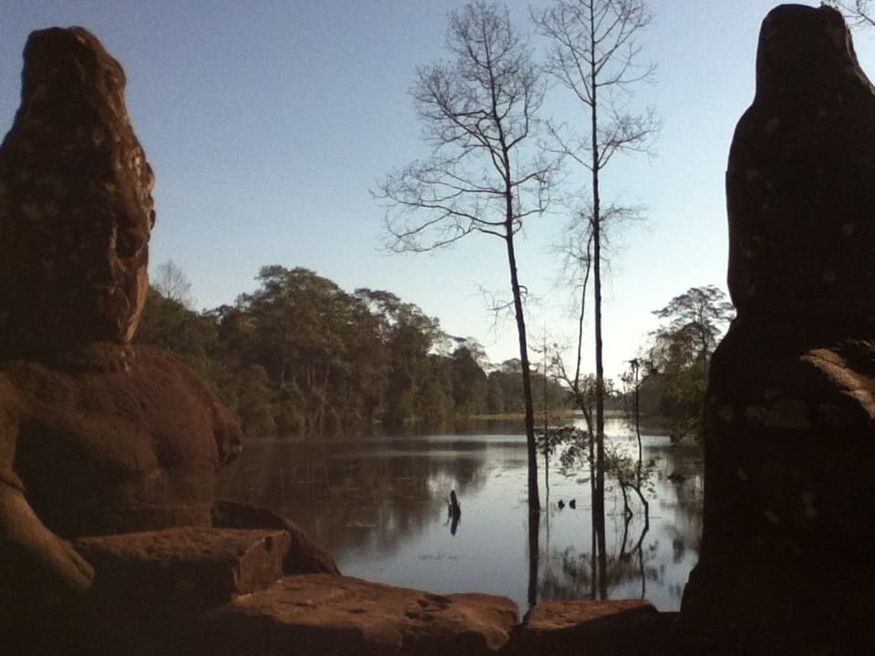 A peaceful scene of a calm lake framed by two weathered stone statues on either side, partially in shadow. Bare trees rise from the water, reflecting softly on the lake's surface, while dense, green forest lines the distant shore under a clear blue sky. The ancient statues, eroded by time, seem to guard the tranquil water, creating a sense of history and serenity in this natural setting.