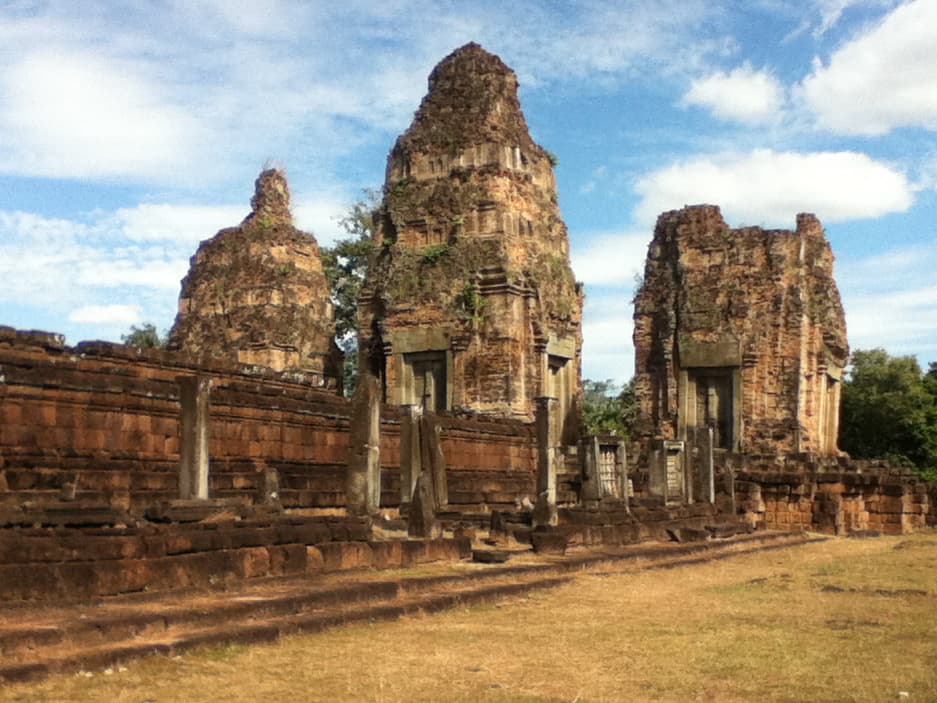 Three ancient, weathered stone towers stand prominently against a blue sky filled with scattered white clouds. The ruins, part of a larger temple complex, show the effects of time, with crumbling edges, uneven surfaces, and overgrown vegetation peeking through the crevices. The reddish-brown stone contrasts with the green surroundings, creating a stark and rugged beauty. In the foreground, a low stone wall with worn pillars leads the viewer’s eye toward the towers, evoking a sense of history and timelessness in the peaceful landscape.
